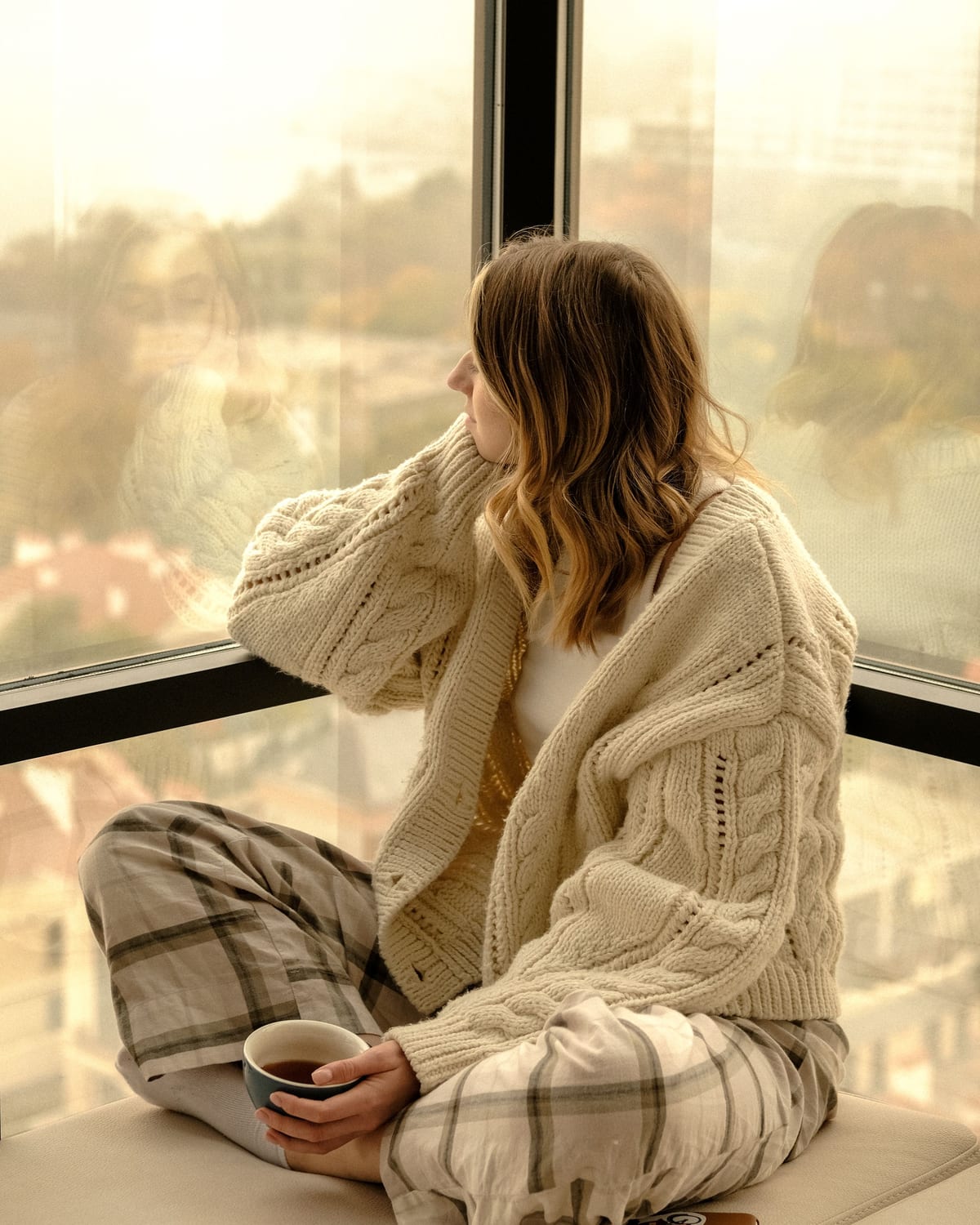 A woman sitting cross-legged on a wide windowsill with a cup of tea, looking out over a city at golden hour